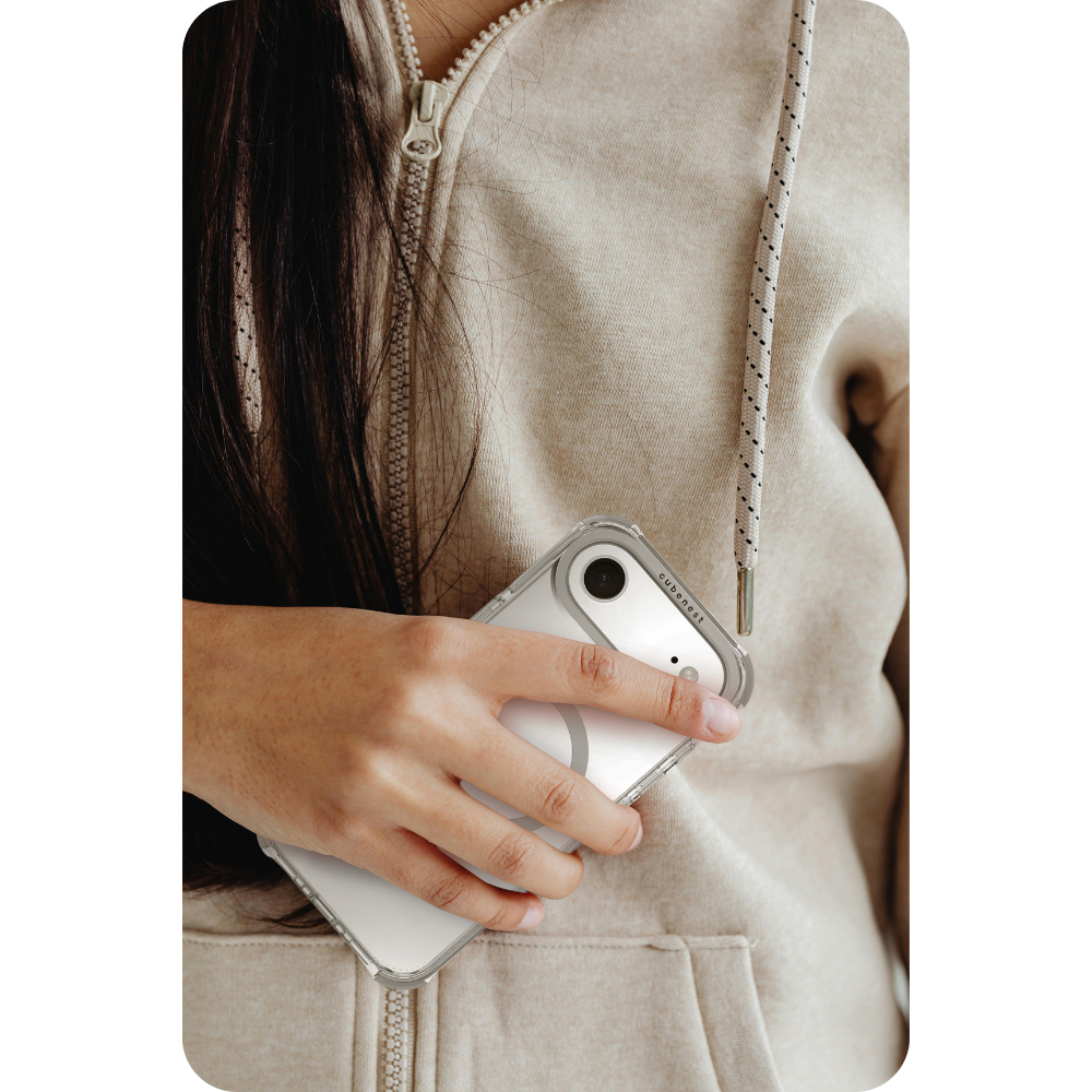 Eine Person hält eine transparente Handyhülle mit einem Riemen. Die Person trägt einen beigen Hoodie. Die Hülle hat eine Kamera und ein weißer Hintergrund.