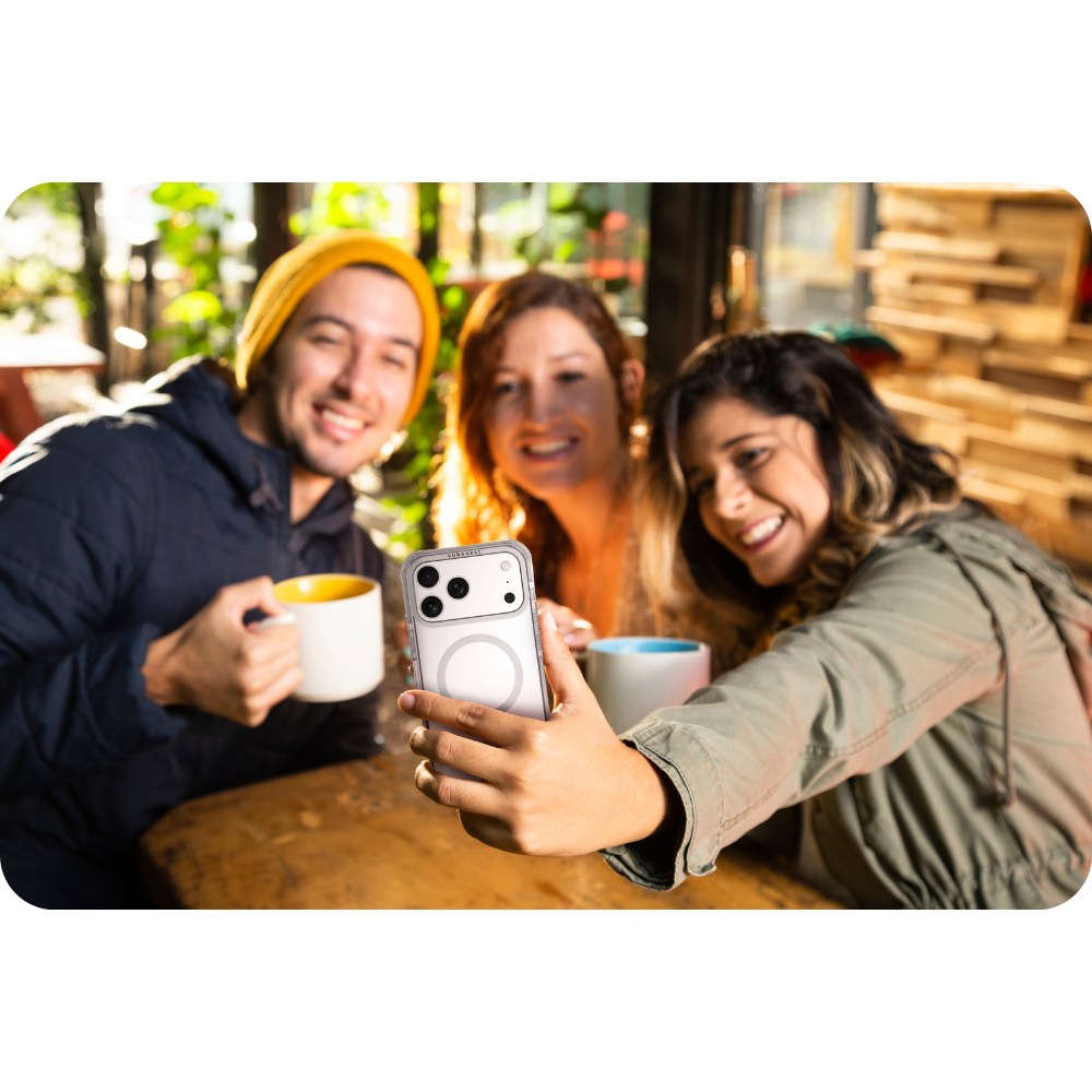 Drei lächelnde Personen machen ein Selfie. Einer hält das Handy. Sie sind draußen in einem Café und halten Kaffeetassen.