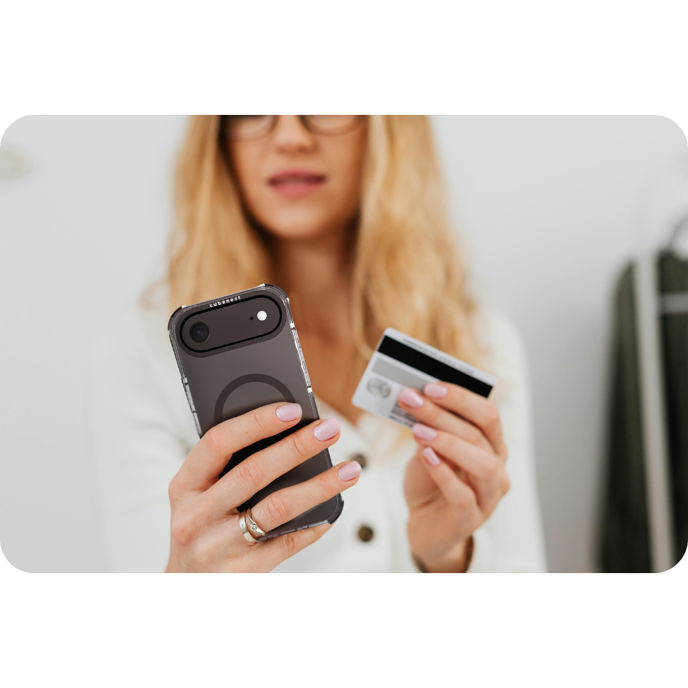Donna che tiene in mano un telefono e una carta di credito. Indossa occhiali e un anello. Il telefono è in una custodia e lo sfondo è sfocato.