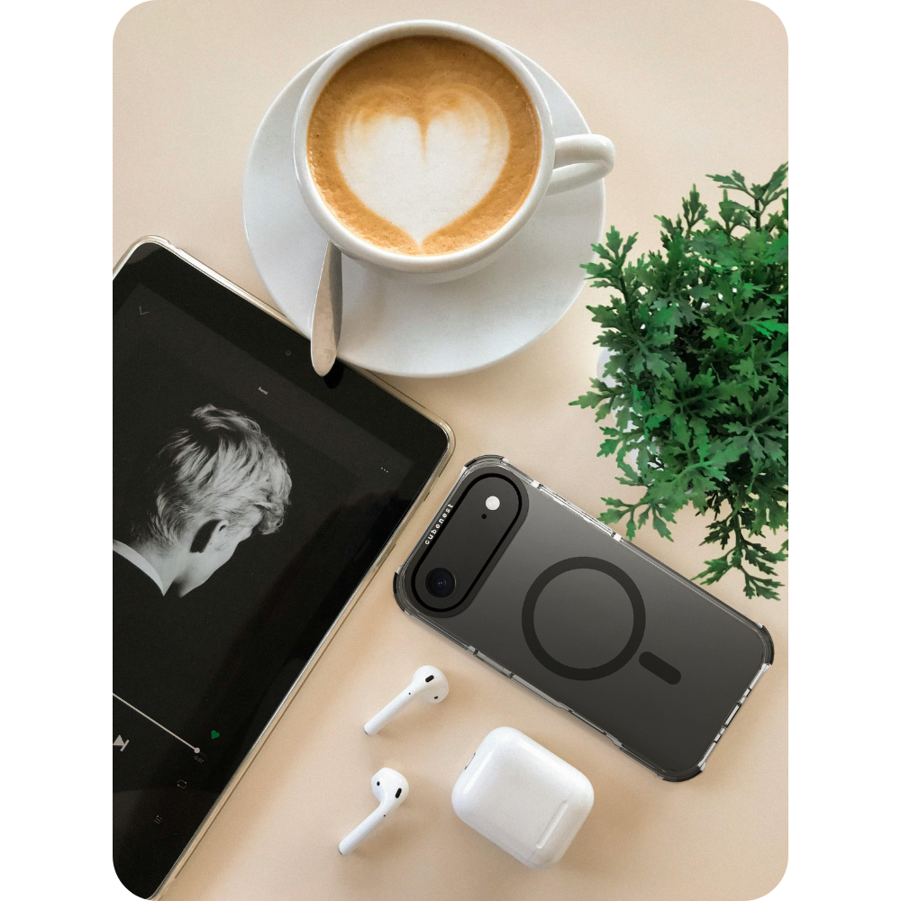 Una tazza di caffè con cuore, un tablet, una custodia per telefono, auricolari e una pianta su una superficie beige.