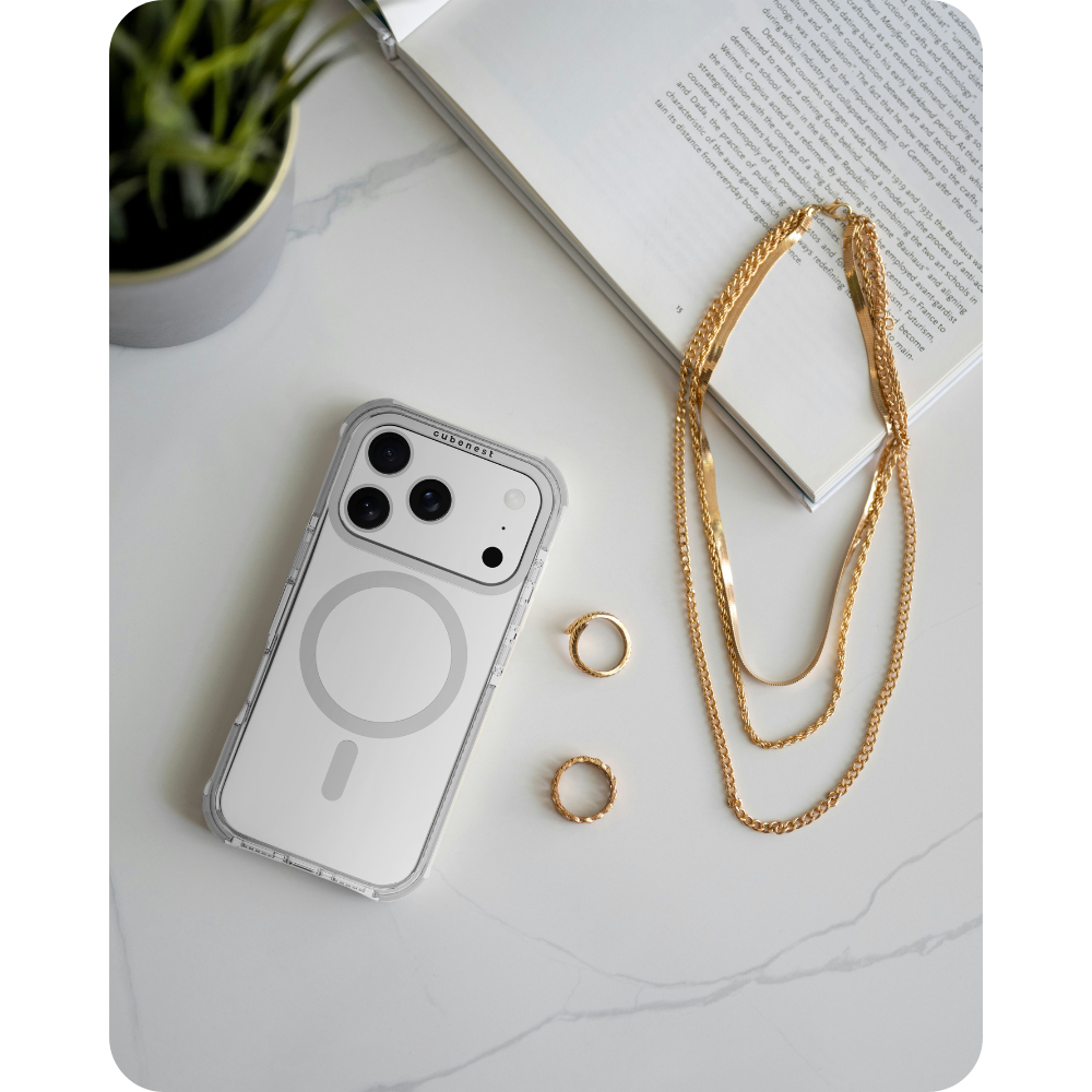 iPhone in klarer Hülle, Goldschmuck und Ringe auf Marmoroberfläche mit Buch und Pflanze.