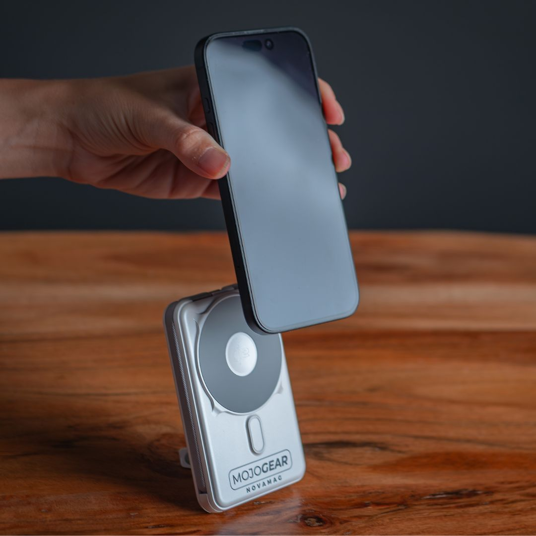 Een hand houdt een zwarte smartphone boven een zilveren MojoGear Novamac-oplader op een houten tafel, donkere achtergrond.