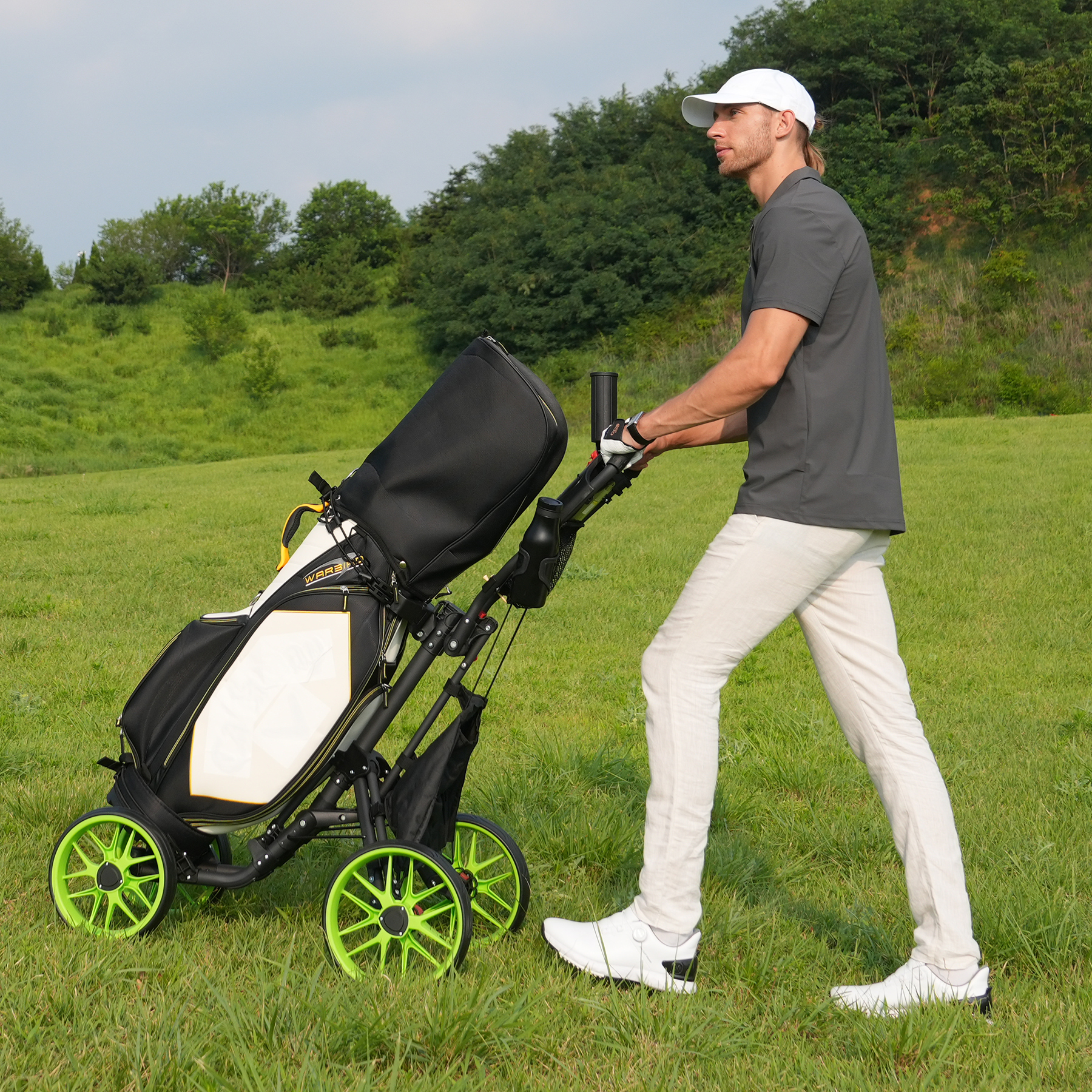 Mężczyzna w białym i szarym stroju golfowym idzie po trawie, ciągnąc wózek golfowy z czarno-białą torbą golfową.