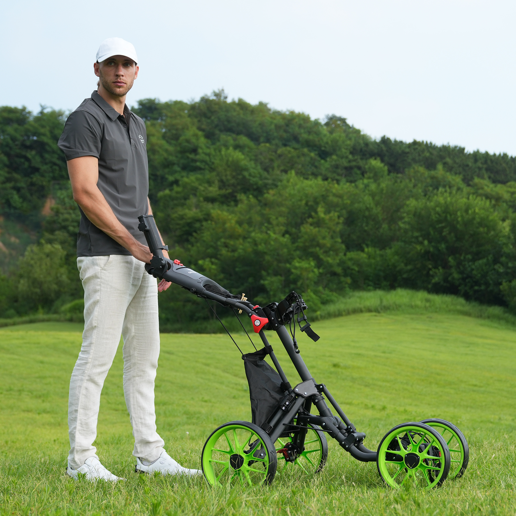 Mężczyzna w czapce i szarej koszulce polo stoi z wózkiem golfowym na zielonym polu. Tło to wzgórze z drzewami.