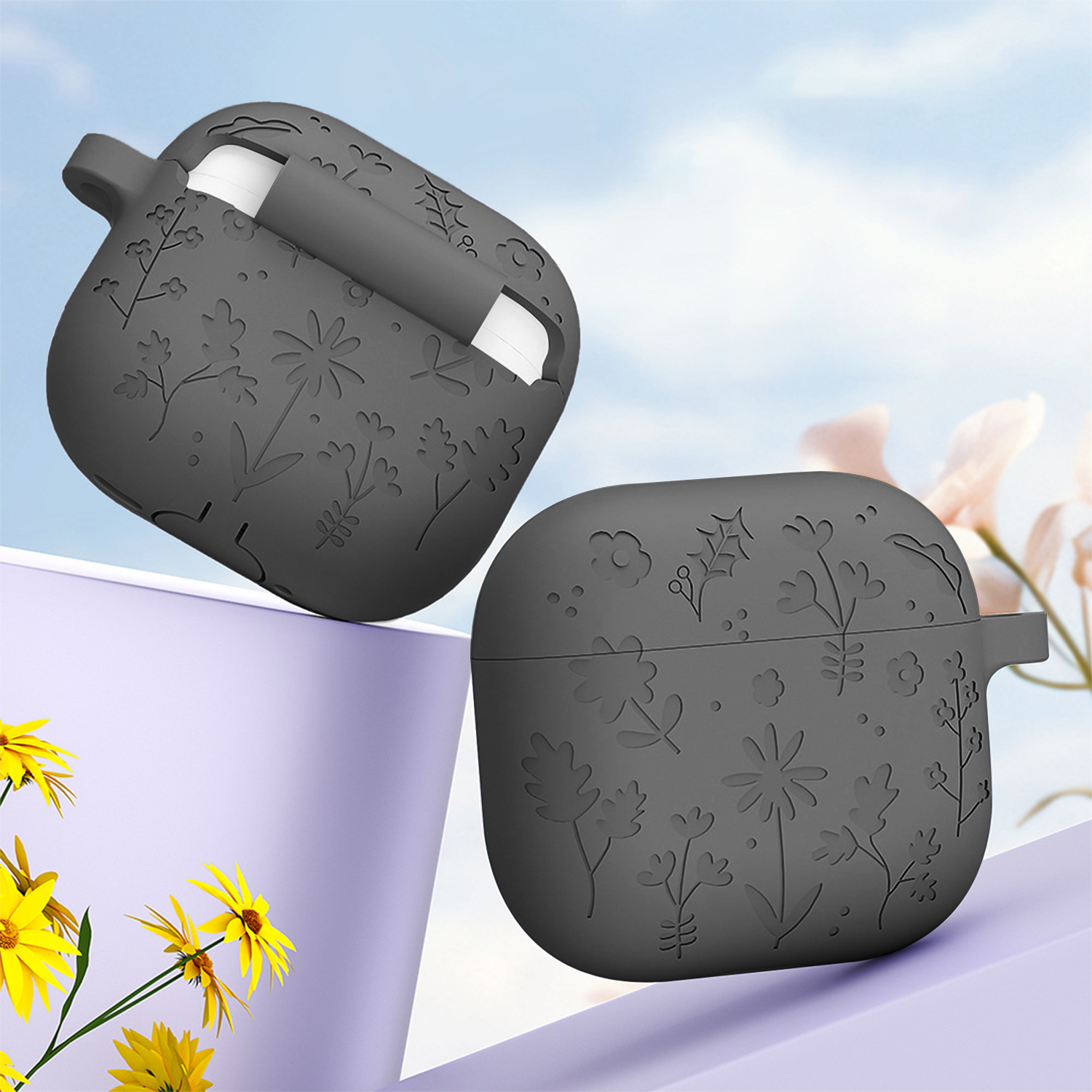 Dos fundas grises para AirPods con patrones florales, una abierta. Flores amarillas y superficie púrpura en primer plano, fondo de cielo.