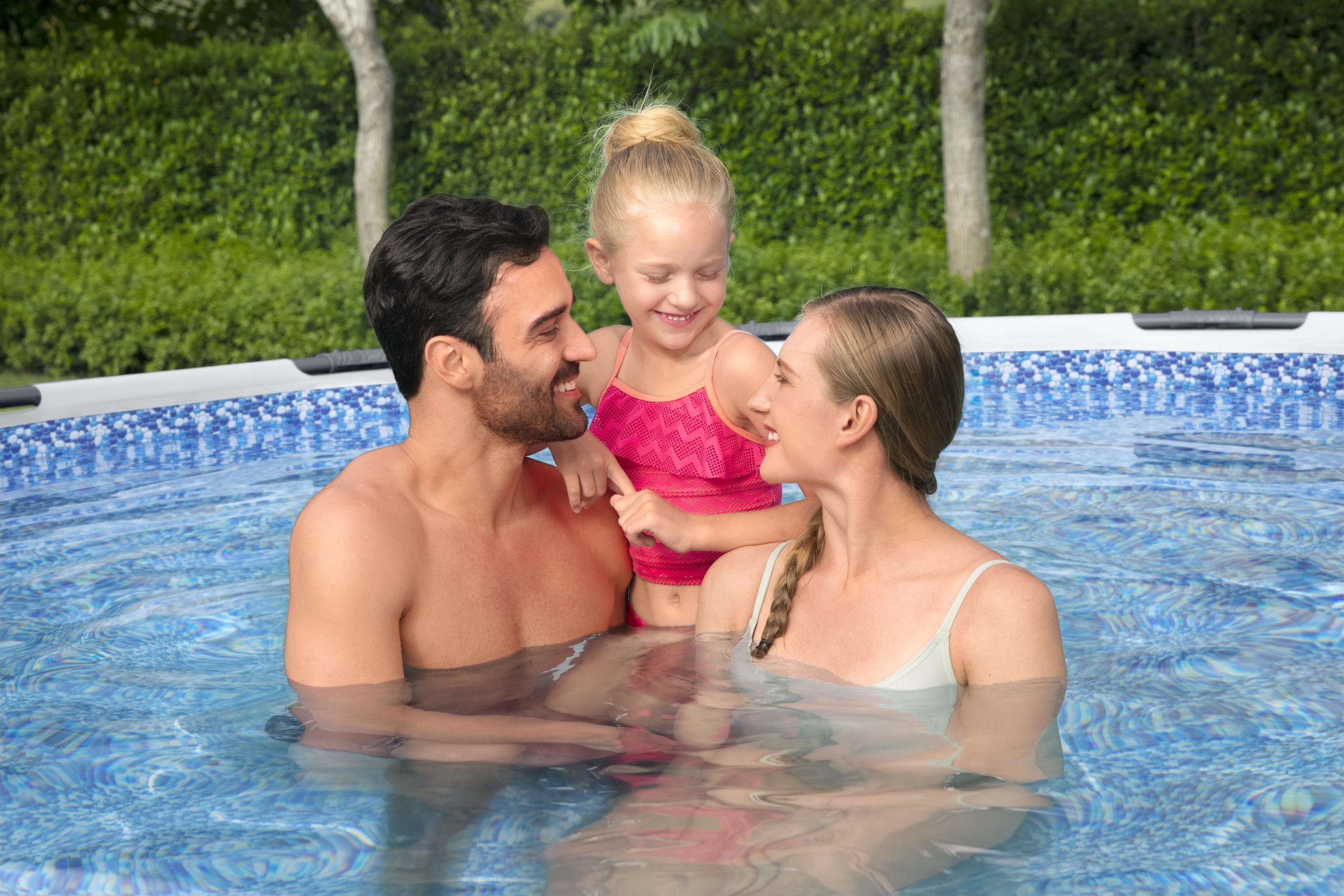 Eine Familie im Pool lächelt sich an. Der Papa und die Mama sind im Wasser. Ein kleines Mädchen steht zwischen ihnen.