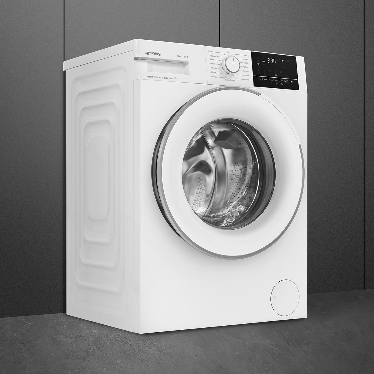 Lavatrice Smeg bianca con porta rotonda, display digitale e pannello di controllo. Si trova su un pavimento grigio contro un muro grigio.