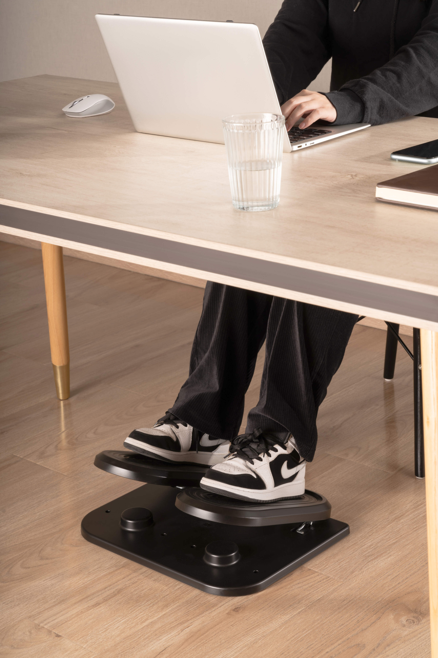 Person typing on laptop at a desk. Feet on a footrest, wearing black and white sneakers. Glass of water and mouse are on the desk.