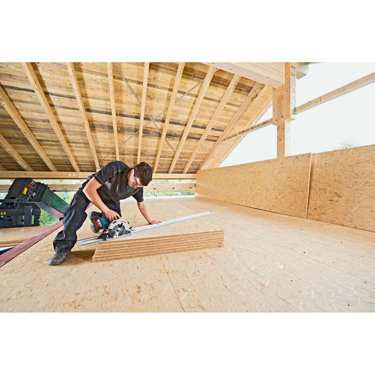 A worker uses a circular saw to cut wooden boards on a construction site. The scene includes wooden beams and OSB panels.