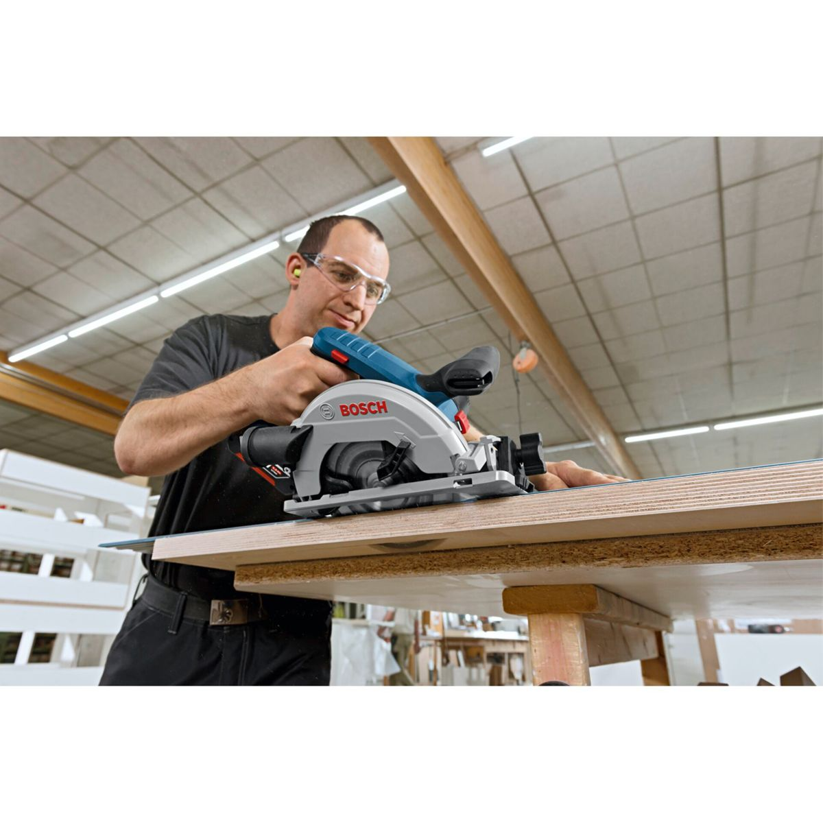 A man using a Bosch circular saw to cut wood. He wears safety glasses and ear protection. The saw is on a wooden table.