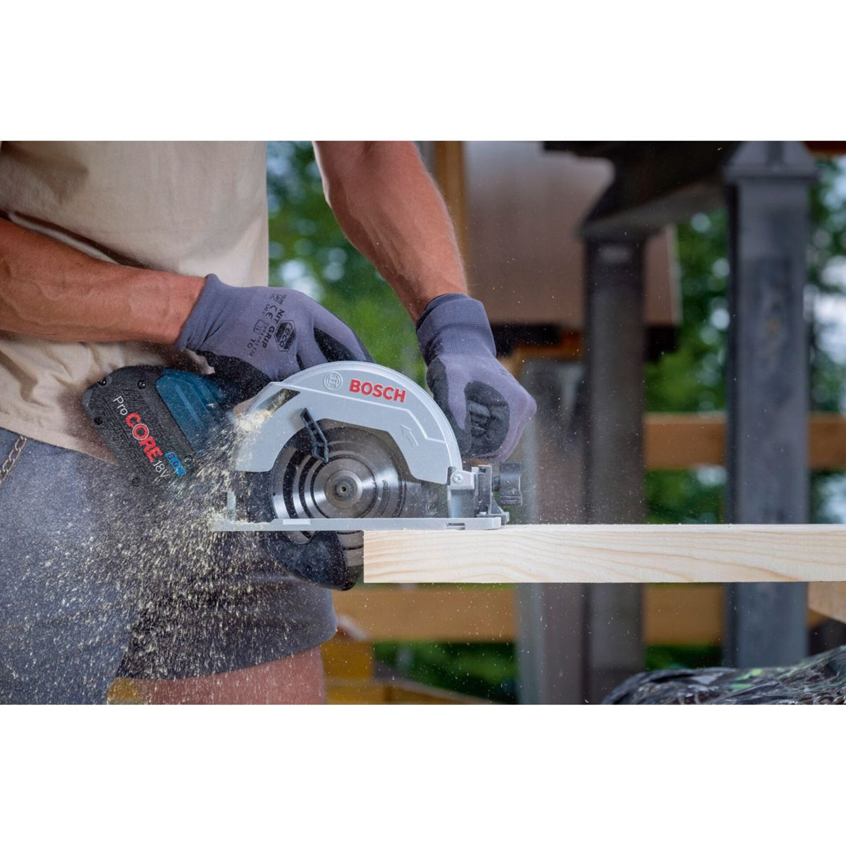 A person is using a circular saw to cut a wooden plank. The saw is grey and blue, with the Bosch logo. Sawdust is flying.