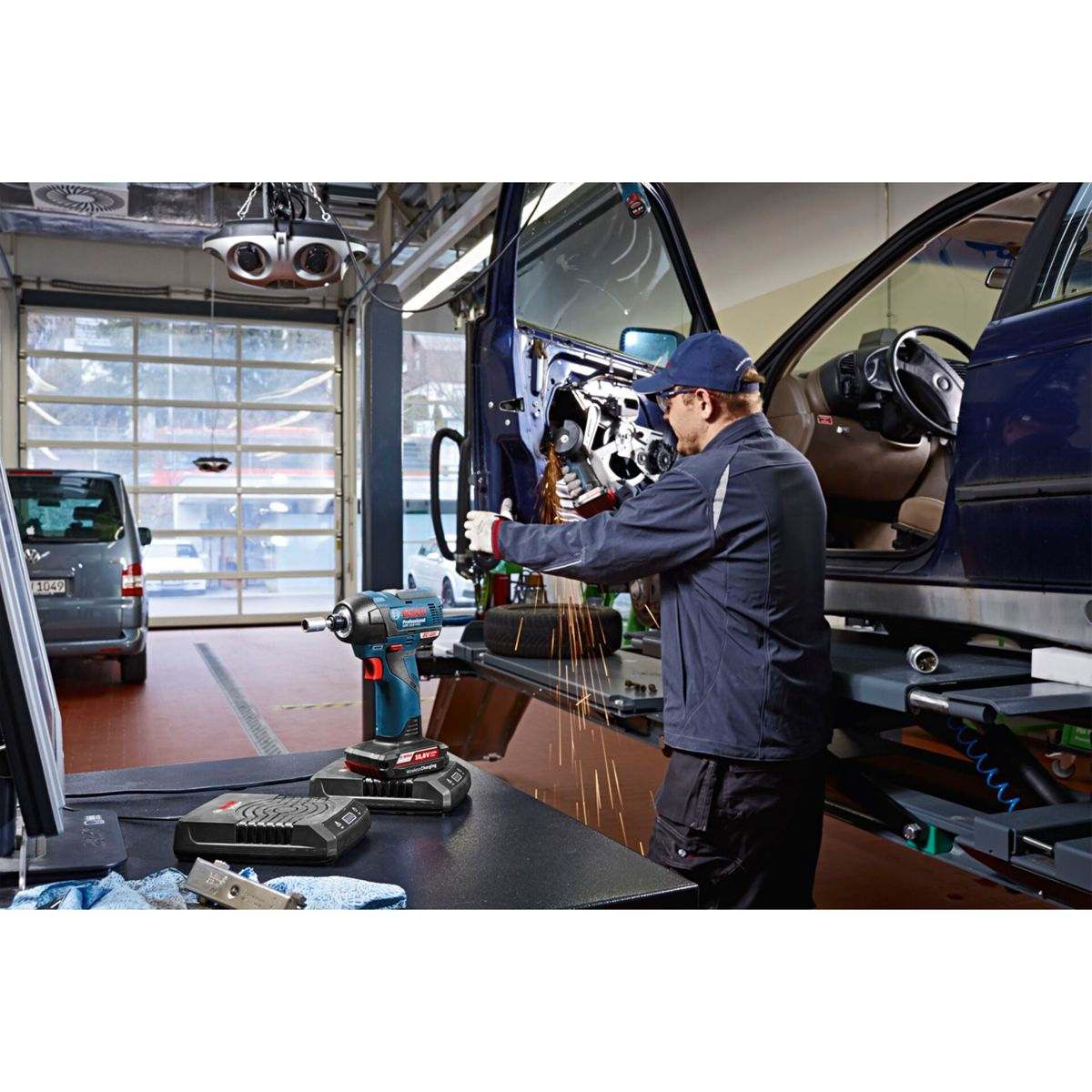 Mechanik w warsztacie używa szlifierki na drzwiach samochodu. Iskry lecą. Narzędzia i furgonetka widoczne w tle.