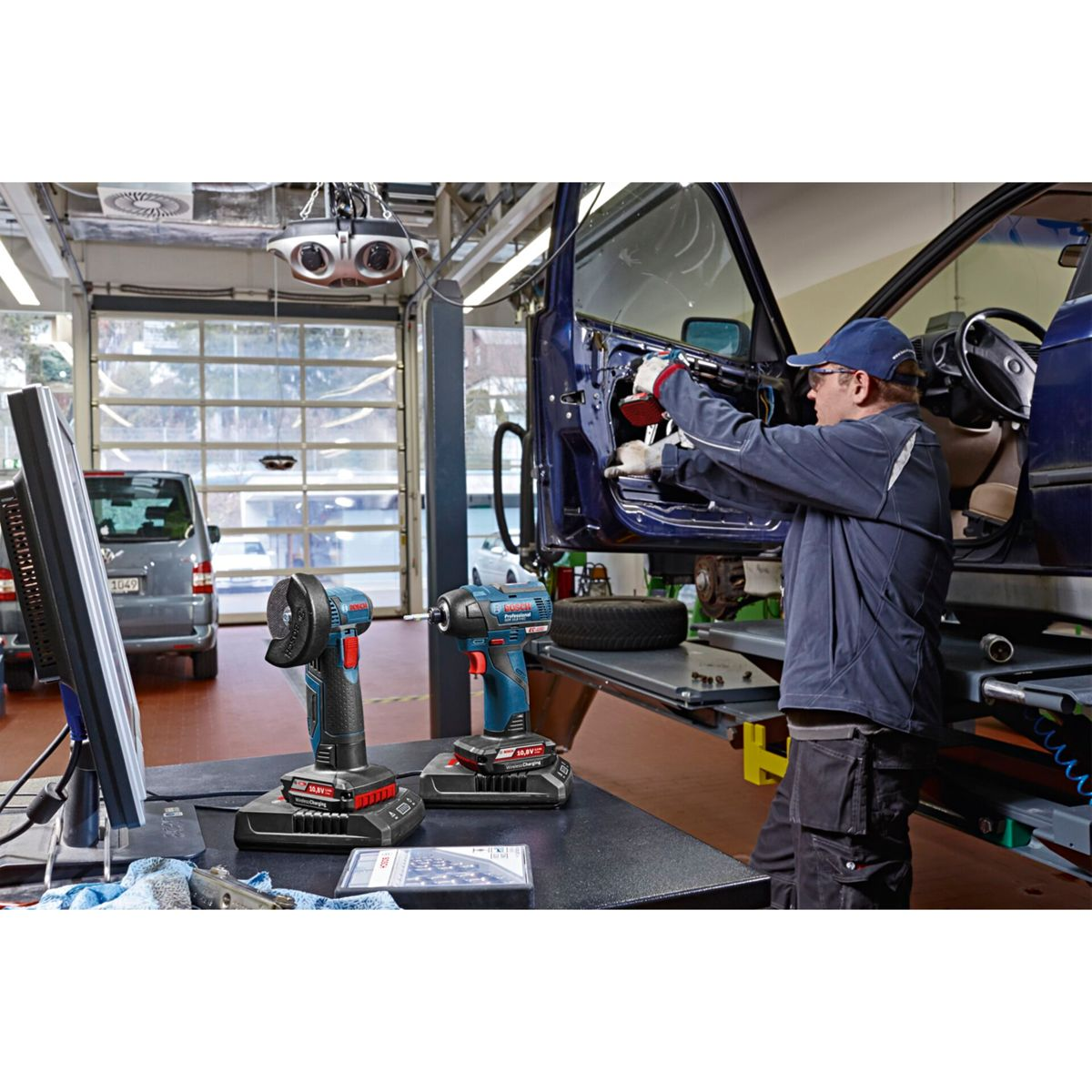 Mechanik pracuje nad samochodem w warsztacie. Elektronarzędzia na stole. Drzwi samochodu otwarte. Szara furgonetka widoczna przez okno.