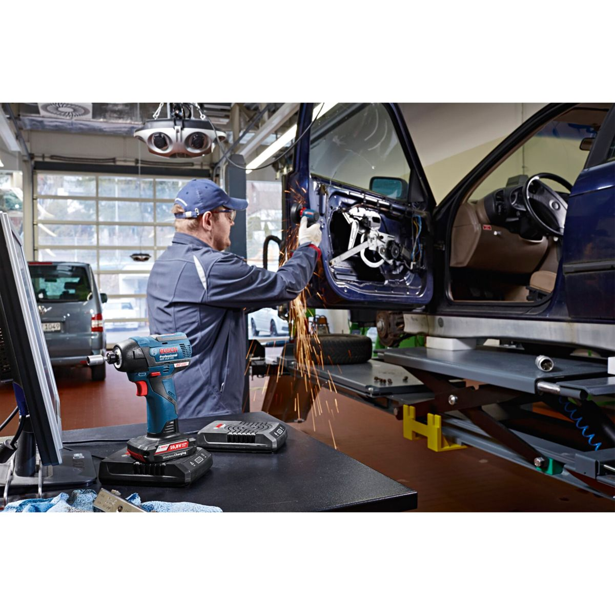 Mechanik spawa drzwi samochodu. Iskry lecą podczas pracy. Narzędzie Bosch leży na stole. W tle widać furgonetkę.