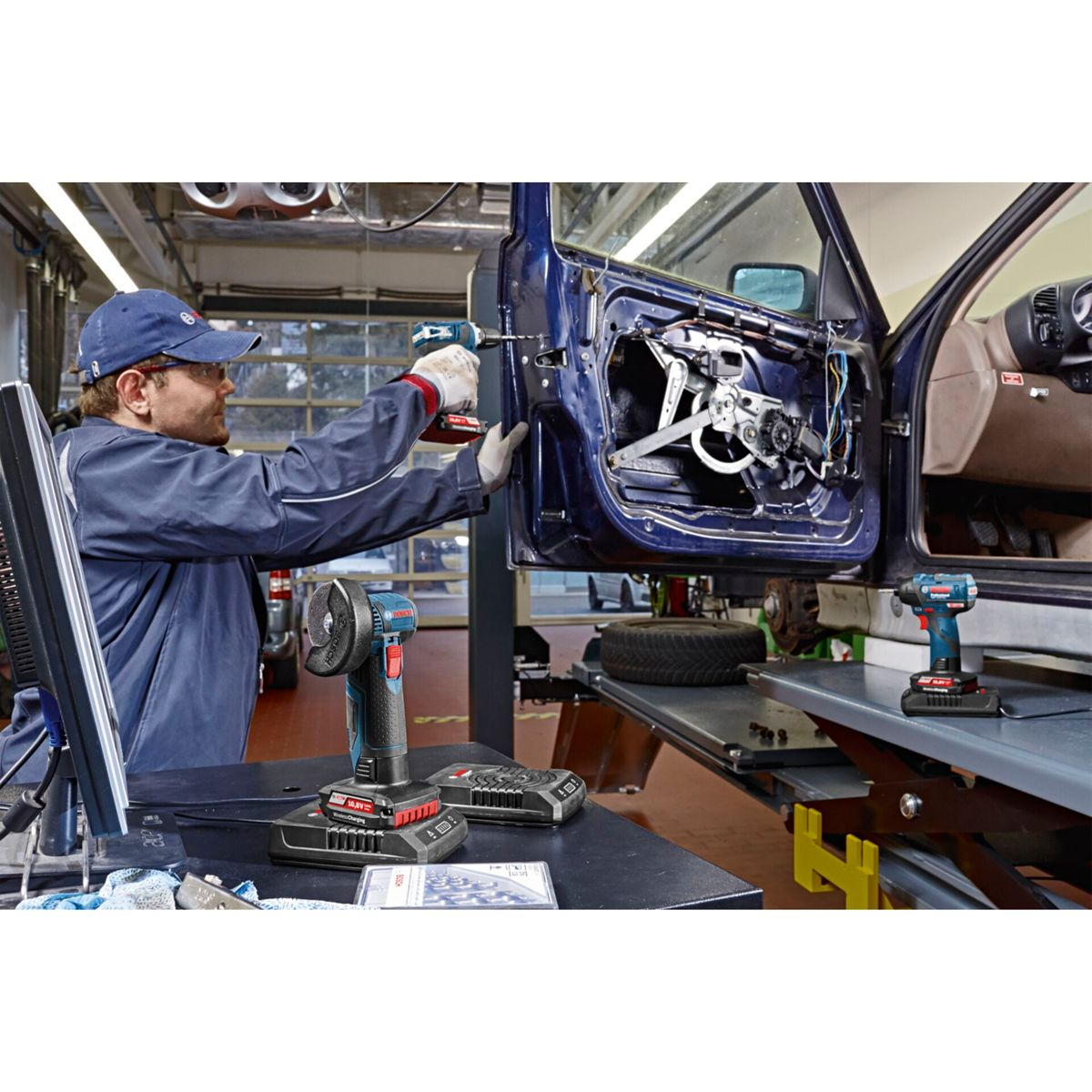 Mechanik w niebieskim kombinezonie używa wiertarki do drzwi samochodu w garażu. Widoczne są narzędzia i części samochodowe.