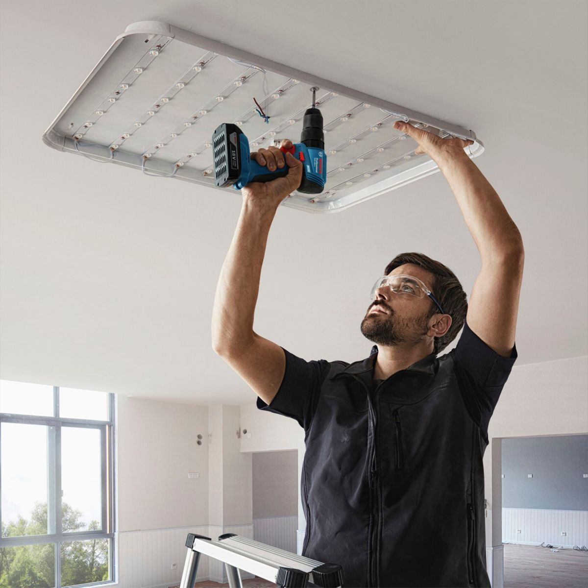 Un uomo installa un apparecchio di illuminazione con un trapano. Indossa occhiali protettivi e si trova sotto l'apparecchio.