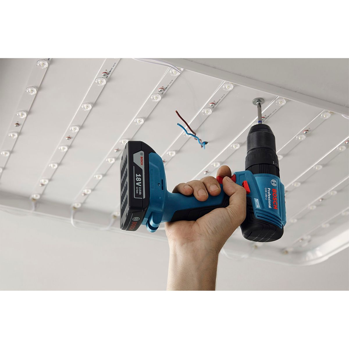 Una mano tiene un trapano blu Bosch, avvitando un pannello a soffitto con luci a LED. I fili sono visibili. Lo sfondo è bianco.