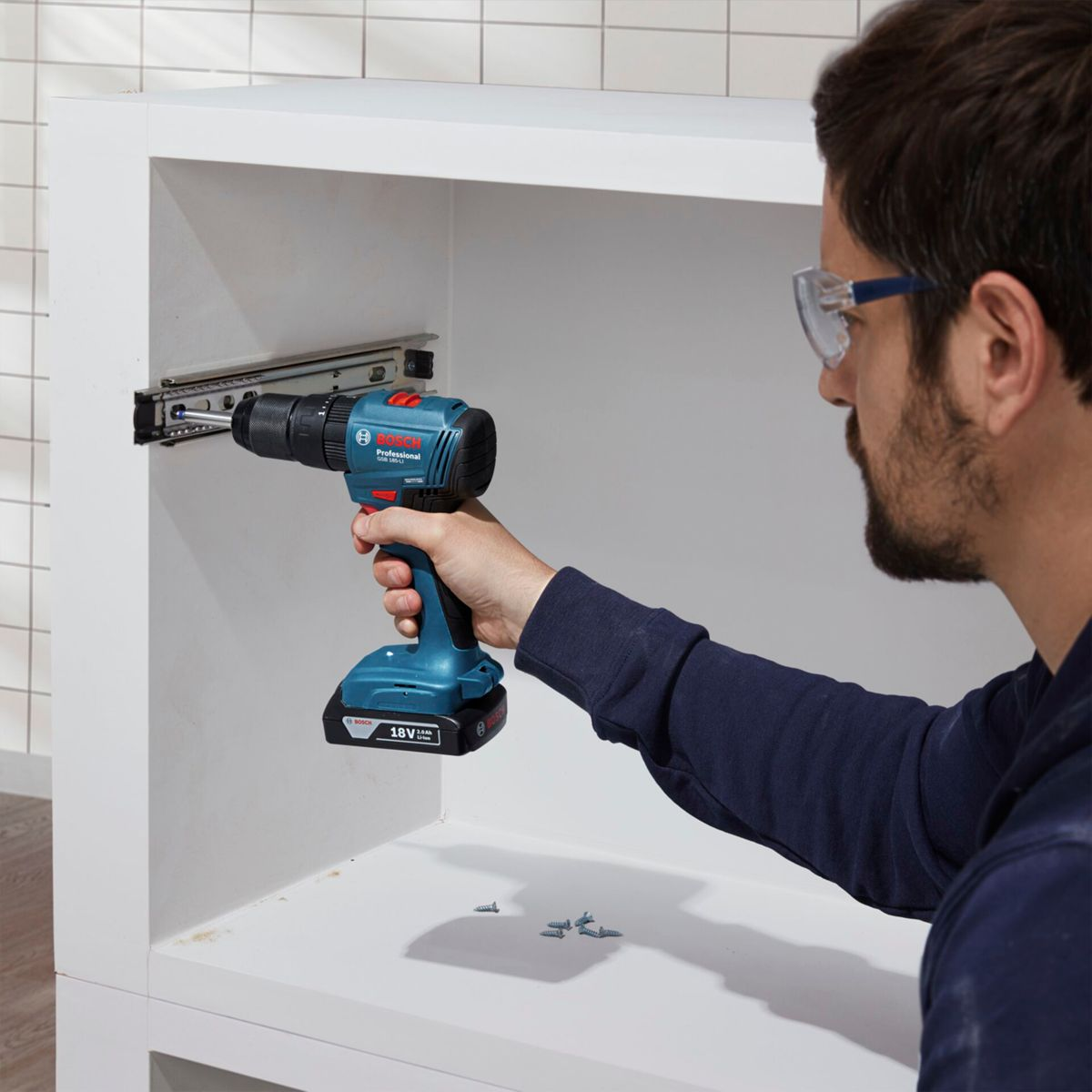Un uomo usa un trapano blu per installare una guida per cassetti in un mobile bianco. Viti sparse.
