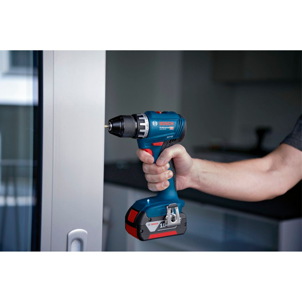 Una mano tiene un trapano Bosch blu e nero. Il trapano è puntato verso una superficie bianca. Lo sfondo mostra un'area cucina.