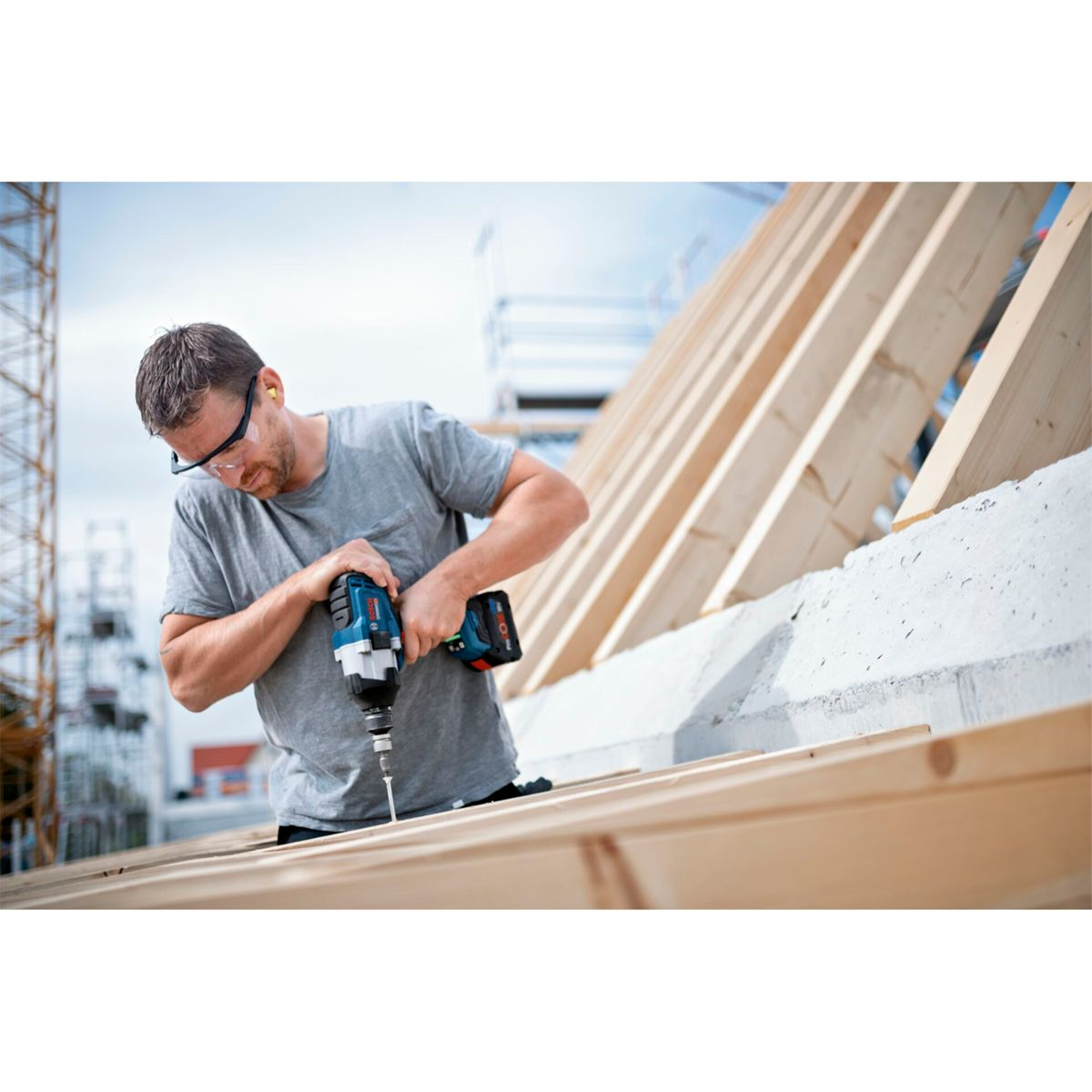 Mężczyzna używa niebiesko-czarnej wiertarki na drewnianych belkach. Nosi okulary i szarą koszulę. Plac budowy w tle.