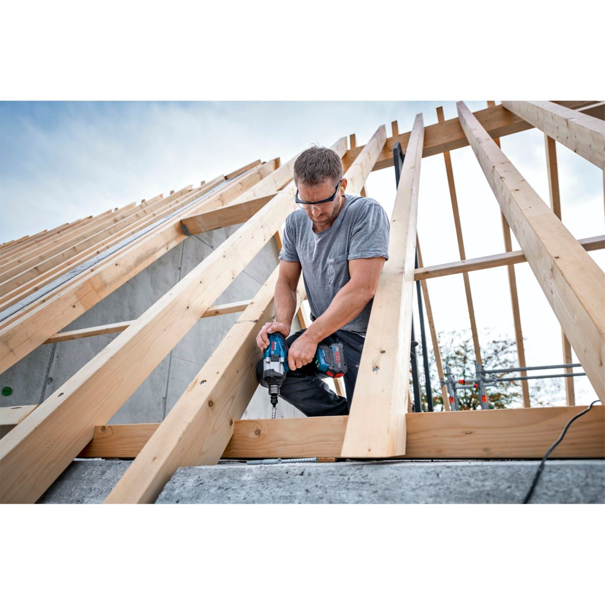 Mężczyzna w szarej koszuli i okularach ochronnych używa wiertarki do drewnianej ramy dachu. Niebo jest niebieskie.