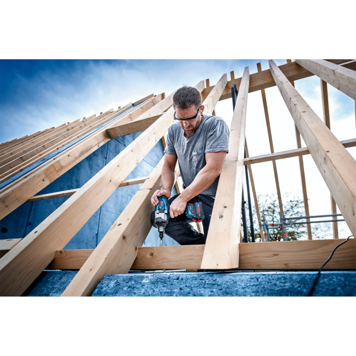 Mężczyzna w szarej koszuli i okularach ochronnych używa wiertarki na konstrukcji dachu. Niebo jest niebieskie.