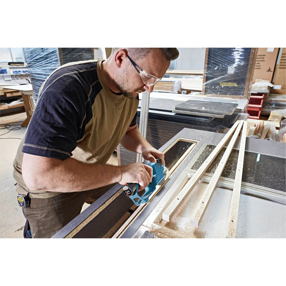 Mężczyzna w okularach ochronnych używa elektronarzędzia do cięcia drewna. Pracuje nad ramą drzwi. Widoczne są trociny.