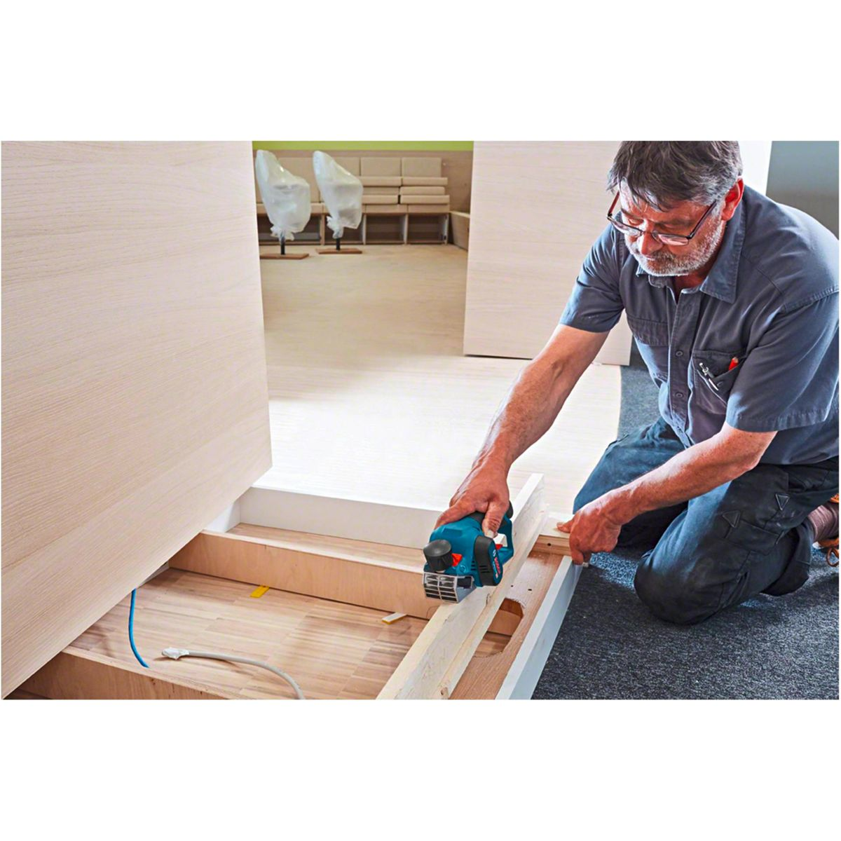 Mężczyzna używa strugarki do ramy drewnianej. Ma okulary i niebieską koszulę. Tło przedstawia pokój z drewnianymi drzwiami.