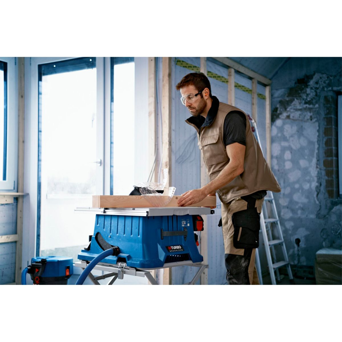 Un uomo in tuta sta tagliando legno con una sega da tavolo blu in un cantiere. Sono visibili anche un aspirapolvere e una scala.