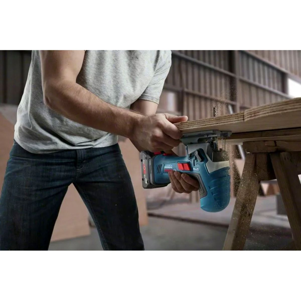 Mężczyzna w dżinsach i szarej koszuli używa niebieskiej piły do cięcia kawałka drewna na drewnianym stole. Wióry lecą.