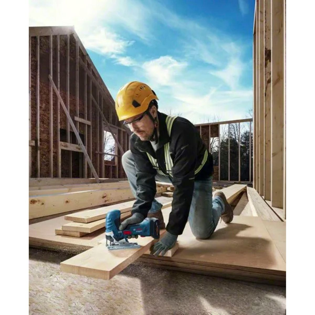 Stolarz tnie drewno wyrzynarką. Ma żółty kask i kamizelkę odblaskową. Tło to plac budowy z błękitnym niebem.