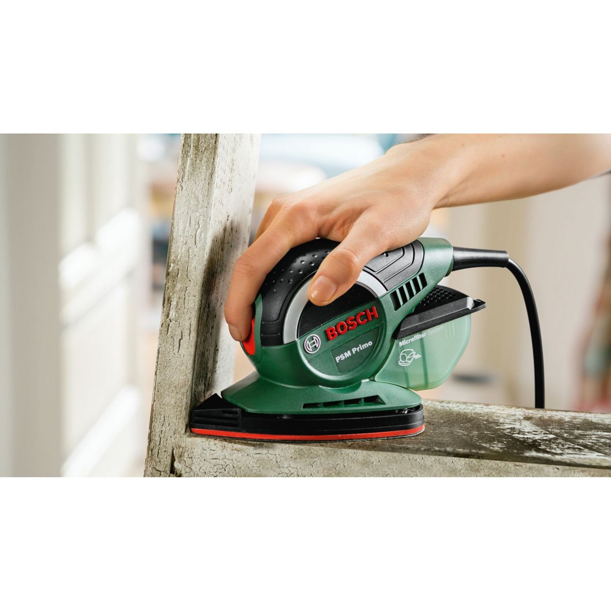 Une main tient une ponceuse Bosch verte, ponçant une surface en bois. La ponceuse est noire et verte avec un accent rouge.