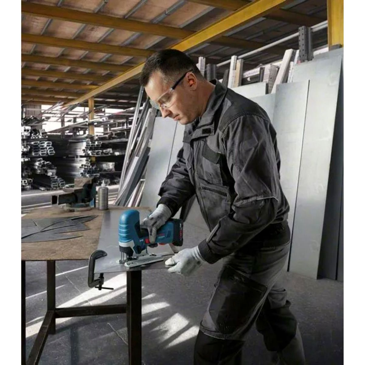 Mężczyzna w okularach ochronnych tnie metal wyrzynarką. Ma na sobie rękawice i kombinezon roboczy. W tle arkusze metalu i półki.