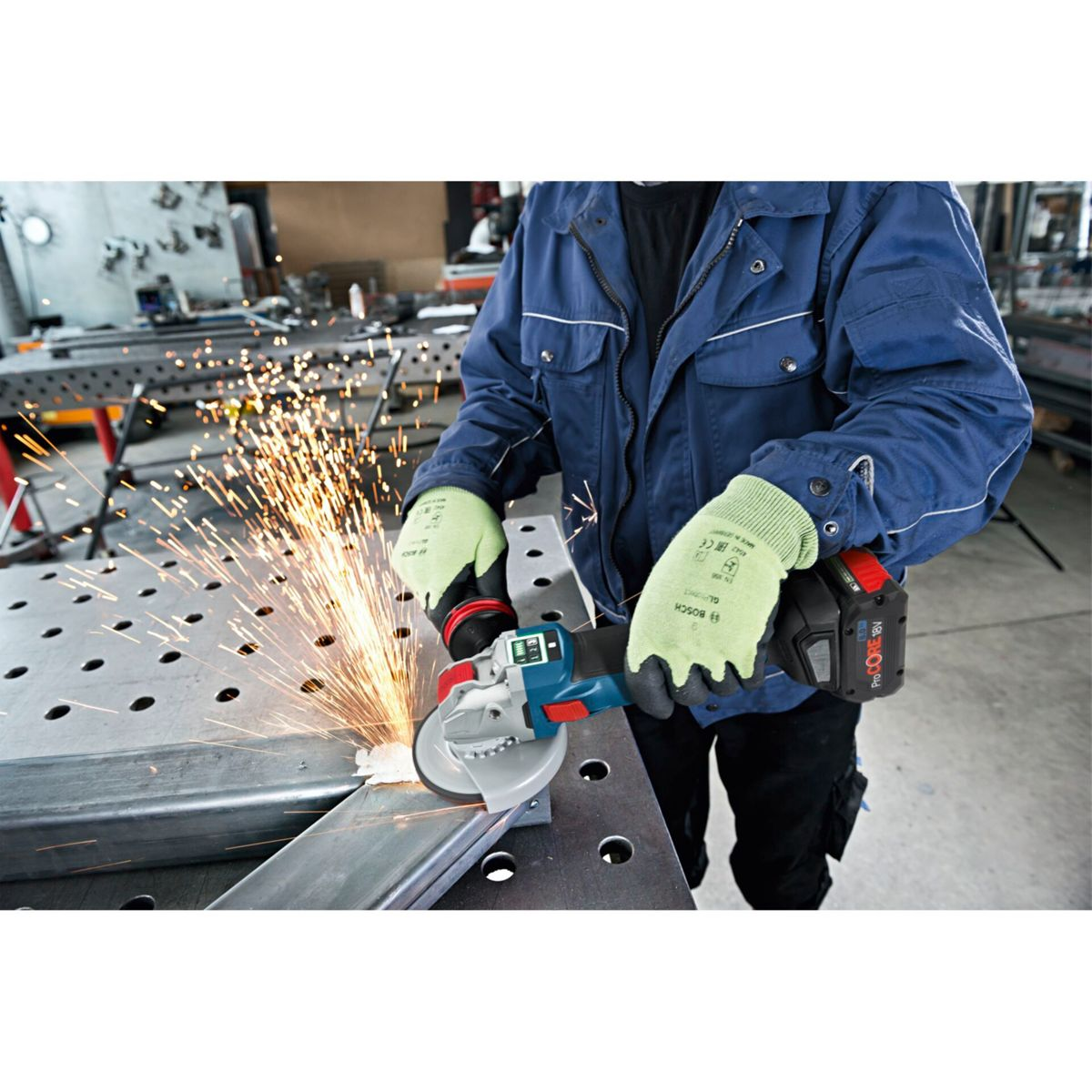 Ein Arbeiter schleift Metall mit einem Winkelschleifer und erzeugt Funken. Er trägt Handschuhe und eine blaue Jacke. Der Hintergrund zeigt eine Werkstatt.
