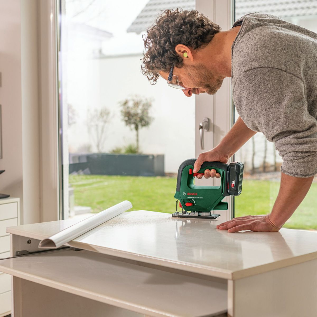 Mężczyzna używa zielonej wyrzynarki na białym stole. Nosi okulary i ochronę słuchu. Widoczne okno i scena zewnętrzna.