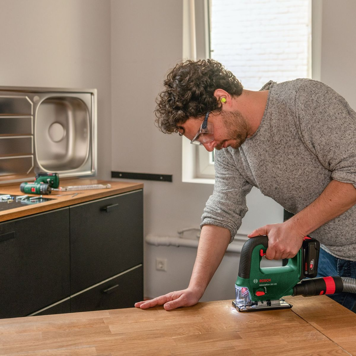 Mężczyzna używa wyrzynarki na drewnianej powierzchni. W tle zlewozmywak, szafki i okno. Ma na sobie okulary ochronne.