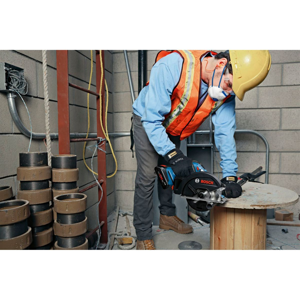 Un operaio edile usa una sega circolare su una bobina di legno. Indossa giubbotto di sicurezza, casco e guanti in un ambiente di cemento.