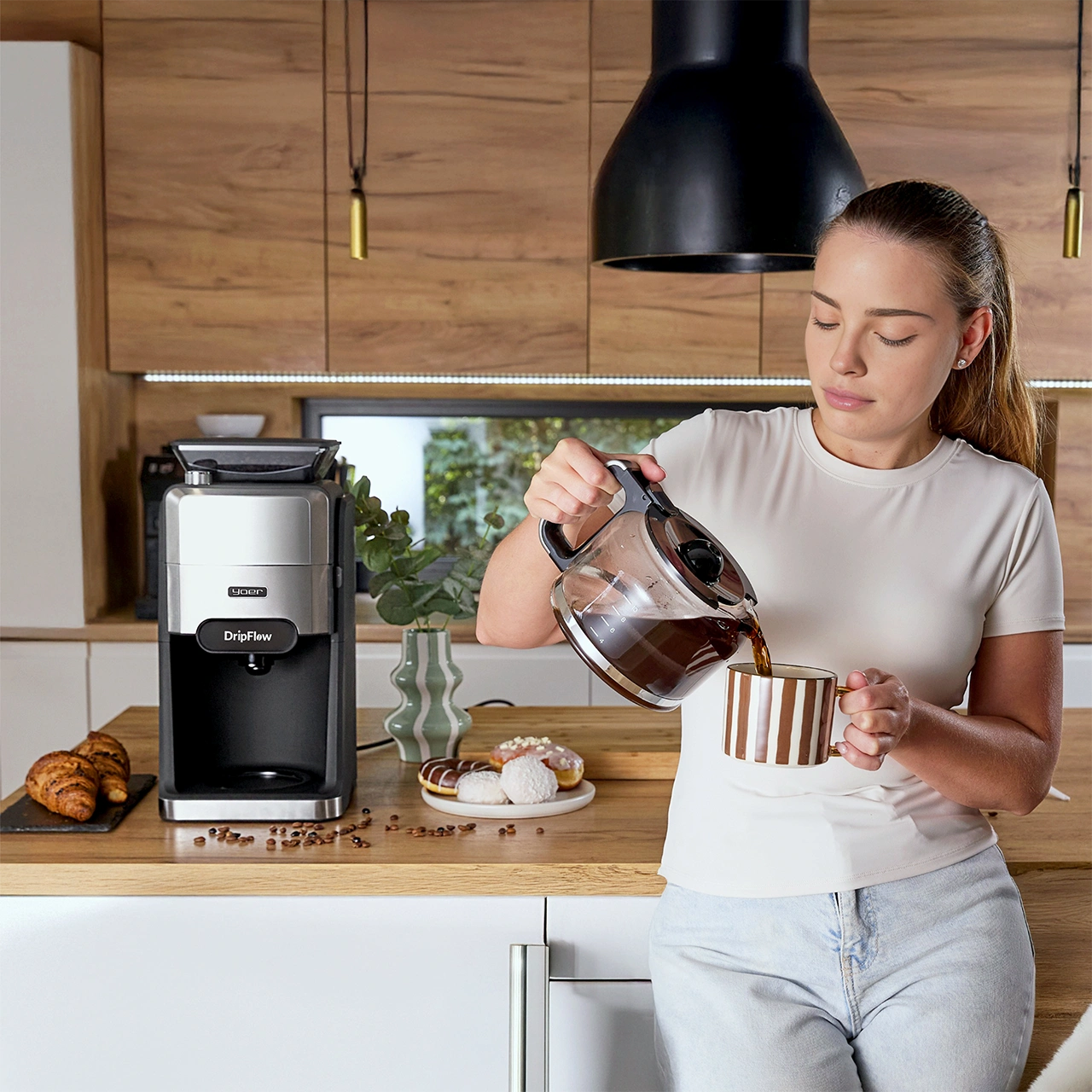 Kobieta nalewa kawę do paskiwanego kubka. Ekspres do kawy i ciastka na blacie. Tło kuchni z drewnianymi szafkami.