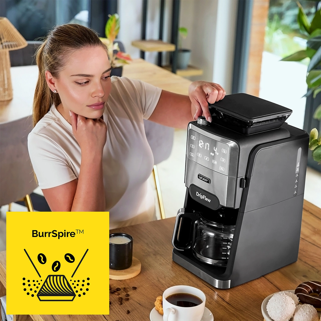 Kobieta używa ekspresu do kawy. Filiżanka kawy, ciastka i logo na drewnianym stole. Zielone rośliny widoczne w tle.