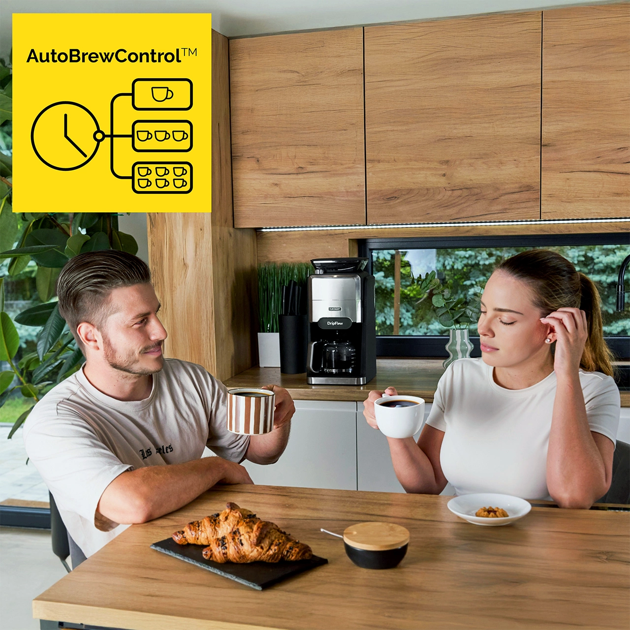 Para siedzi przy stole z kawą i rogalikami. W tle ekspres do kawy. Widoczne logo AutoBrewControl.