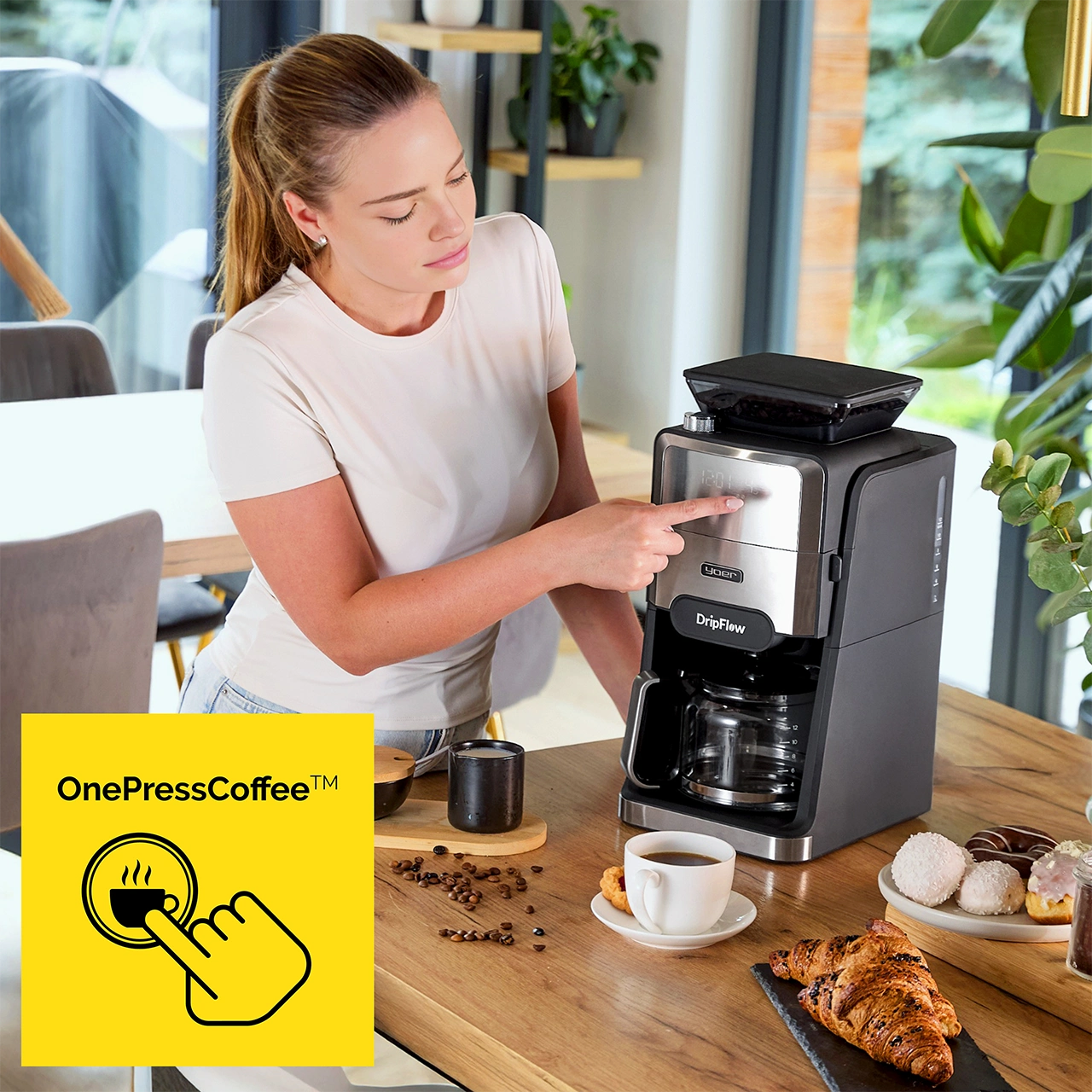 Kobieta obsługuje ekspres do kawy. Filiżanka kawy, ciastka i ziarna na blacie. Widoczne żółte logo OnePressCoffee.
