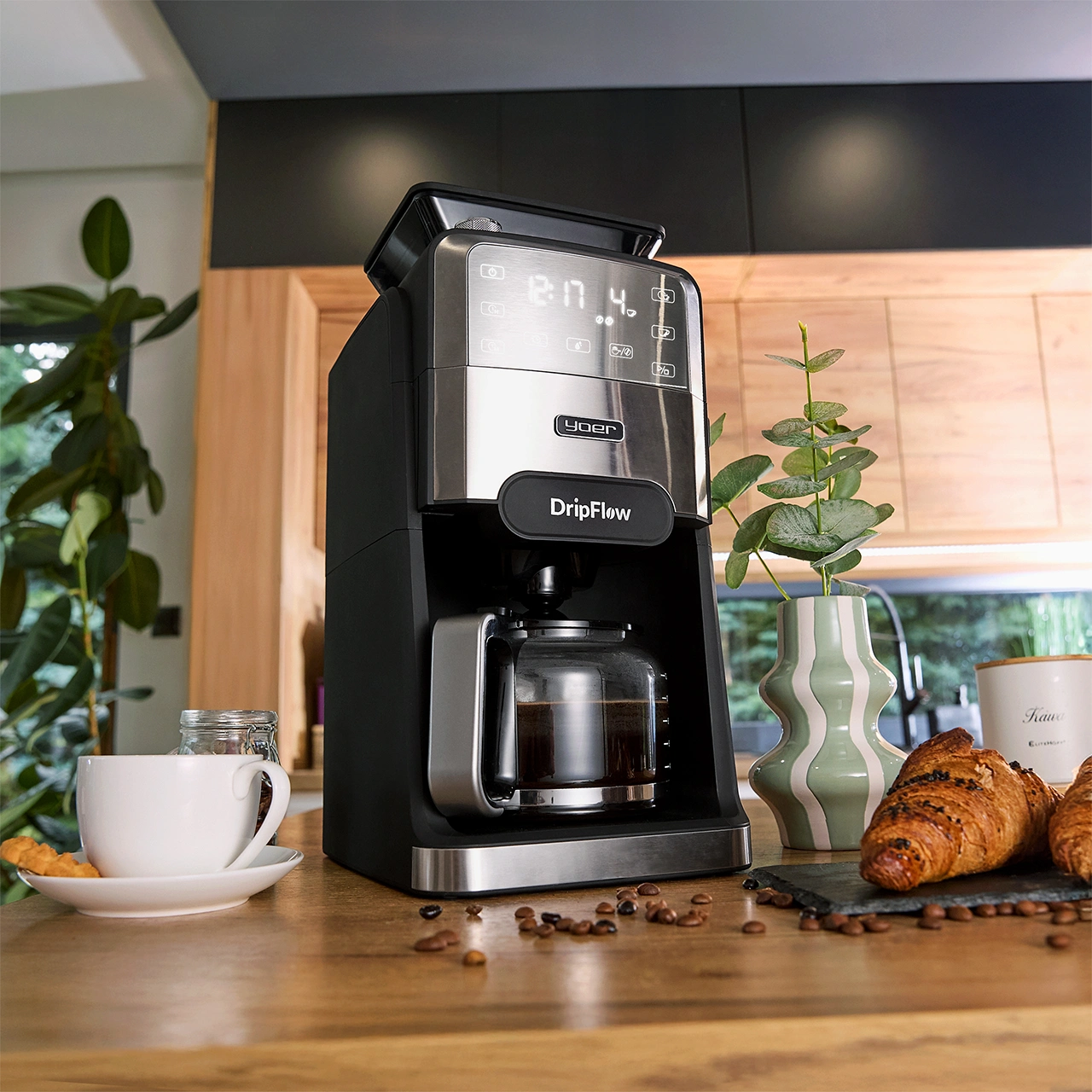 Ekspres do kawy ze szklaną karafką, filiżanką, rogalikiem i wazonem na drewnianym stole. Ziarna są rozrzucone. Tło: kuchnia.