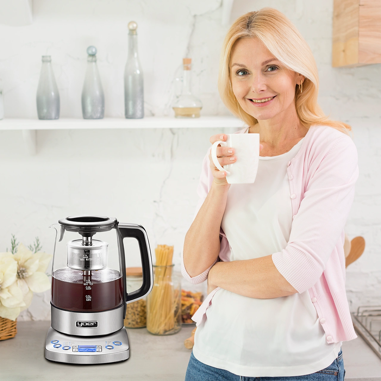 Kobieta w kuchni trzymająca kubek, uśmiechnięta. Czajnik jest na pierwszym planie. Tło ma półki z butelkami.