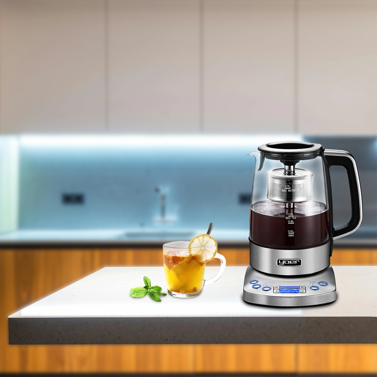 Szklany czajnik z herbatą w środku, obok filiżanki herbaty z cytryną i miętą na białej powierzchni. Tło kuchenne.