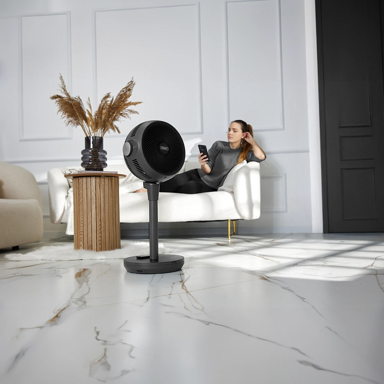Kobieta na sofie z telefonem, wentylator na pierwszym planie. Beżowa sofa, białe ściany, drewniany stół z wazonem, czarne drzwi i marmurowa podłoga.