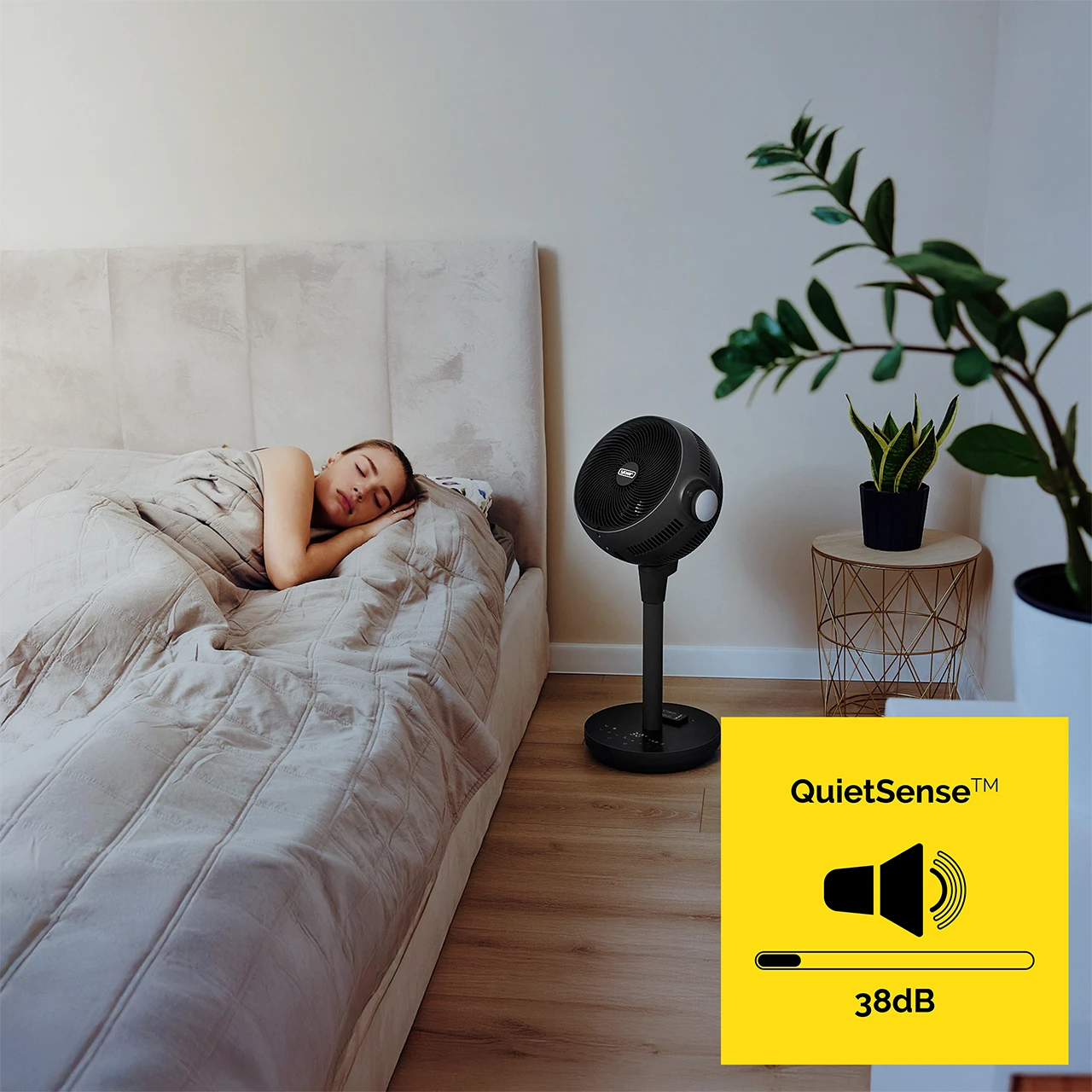 Kobieta śpi w łóżku. Czarny wentylator obok łóżka. Żółty znak wskazuje 'QuietSense' 38dB. Rośliny w tle.