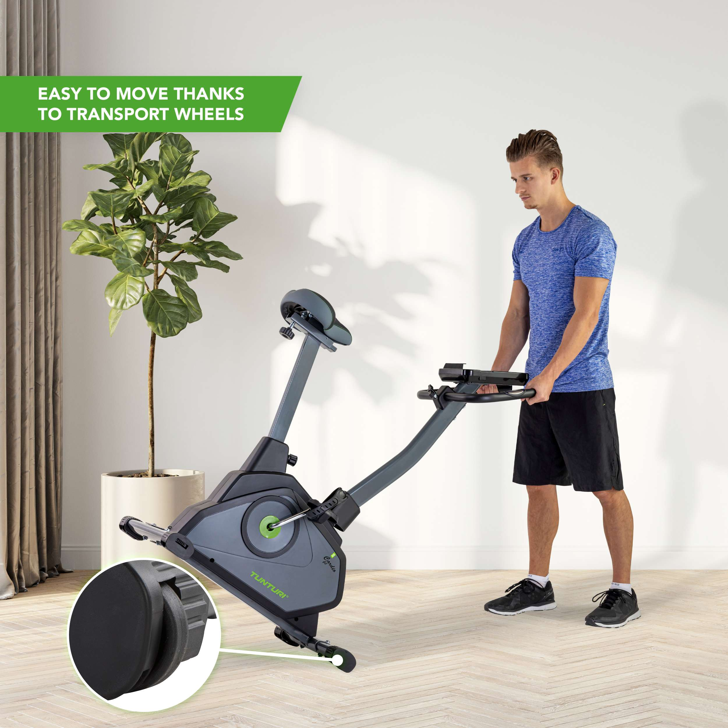 A man moves an exercise bike. It is gray and black. A plant in a white pot is in the background.