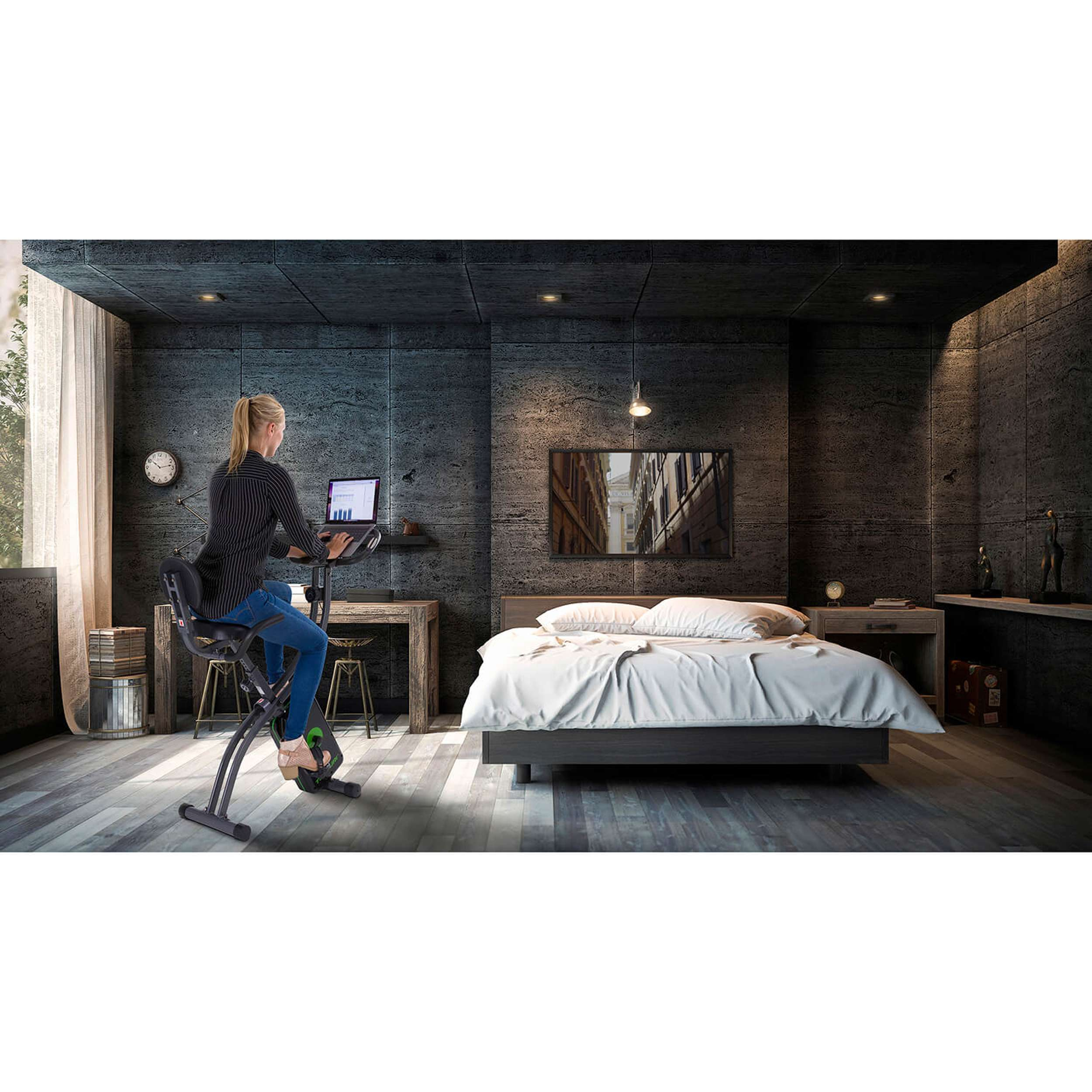 Vrouw op hometrainer in een slaapkamer. Laptop op bureau. Bed met wit beddengoed. Donkergrijze muren en vloer.