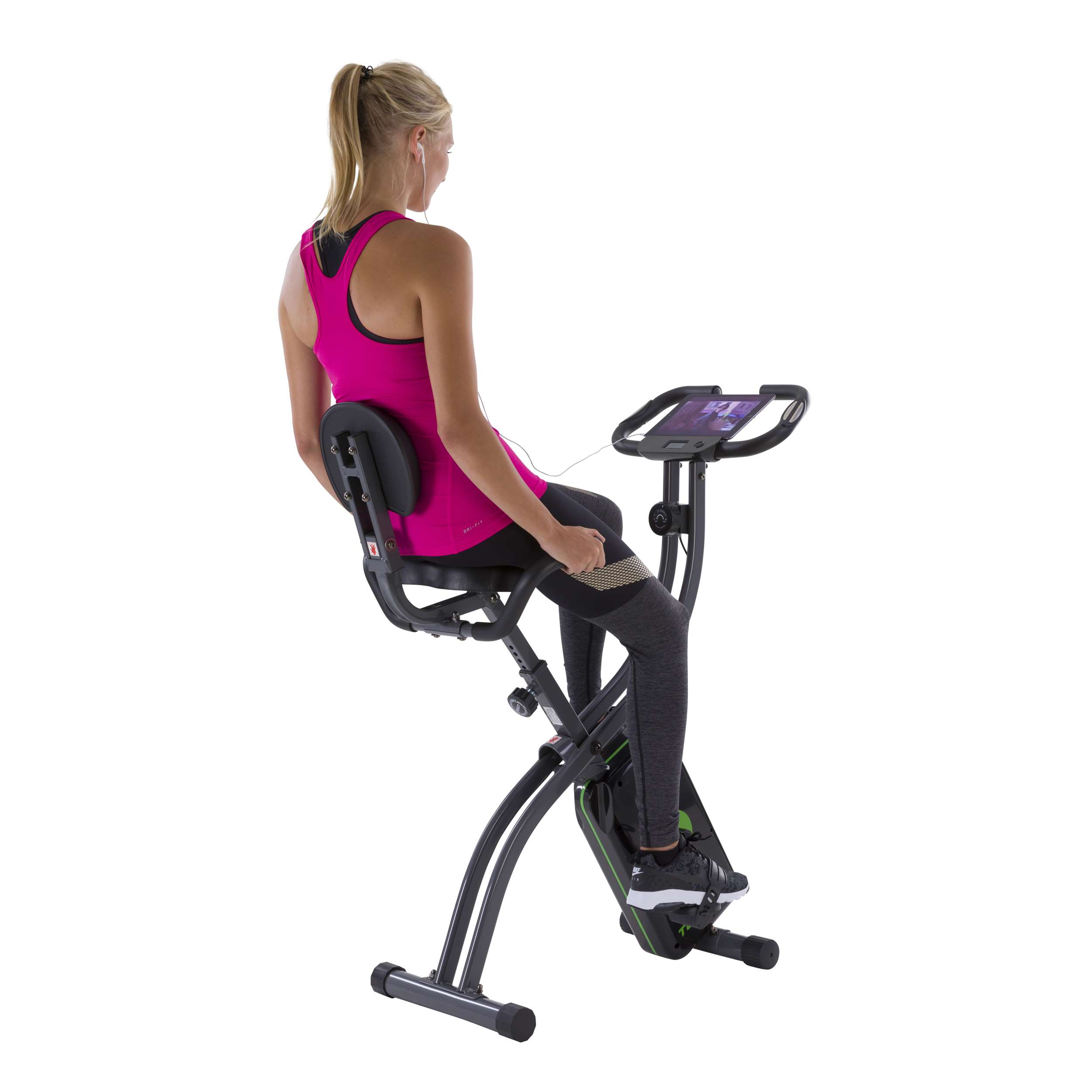 Vrouw op hometrainer. Ze draagt roze top en zwarte legging. Fiets is zwart en groen. Ze heeft oortelefoons.