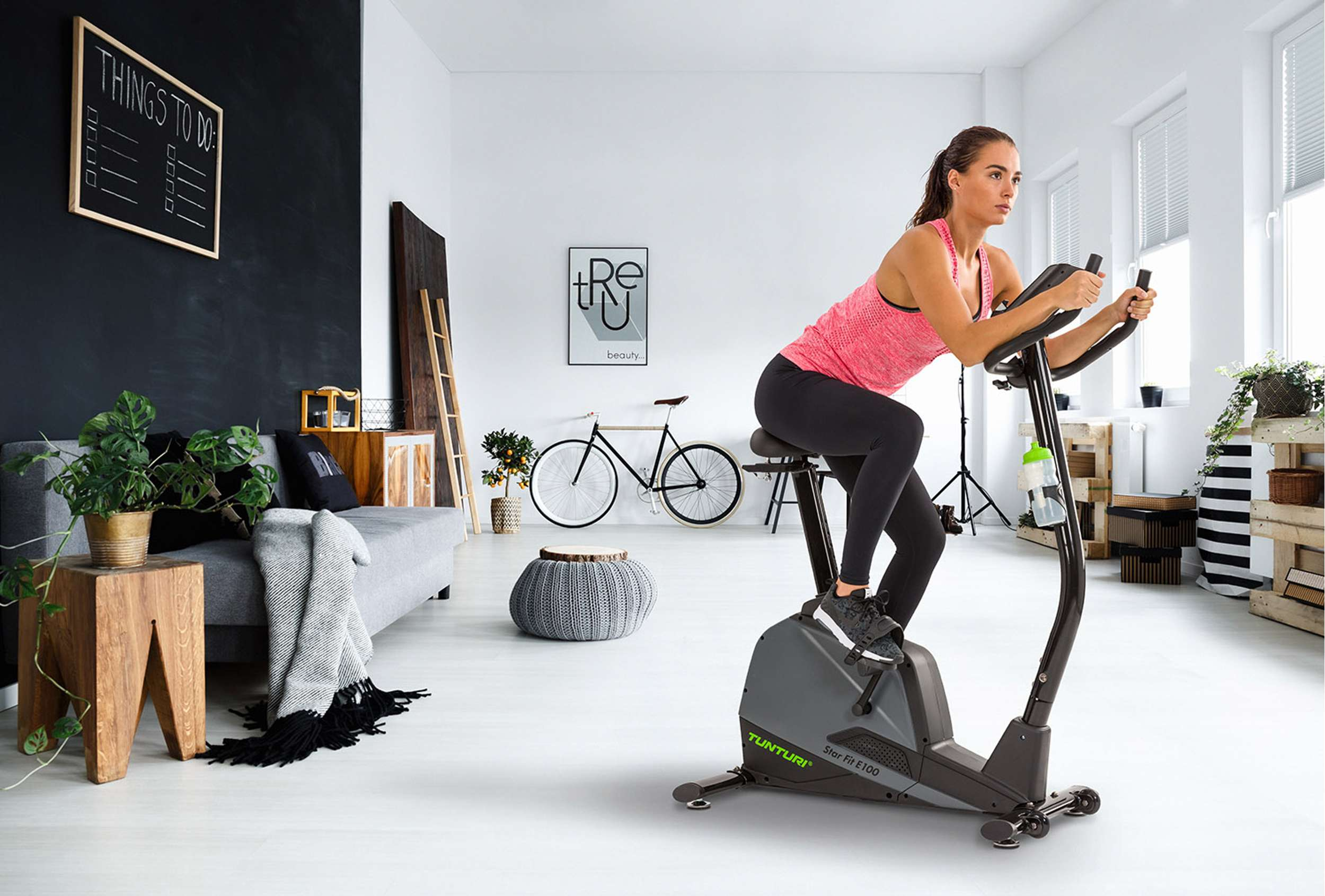 Een vrouw traint op een hometrainer. De kamer is minimalistisch. Een zwart schoolbord met een takenlijst staat links.